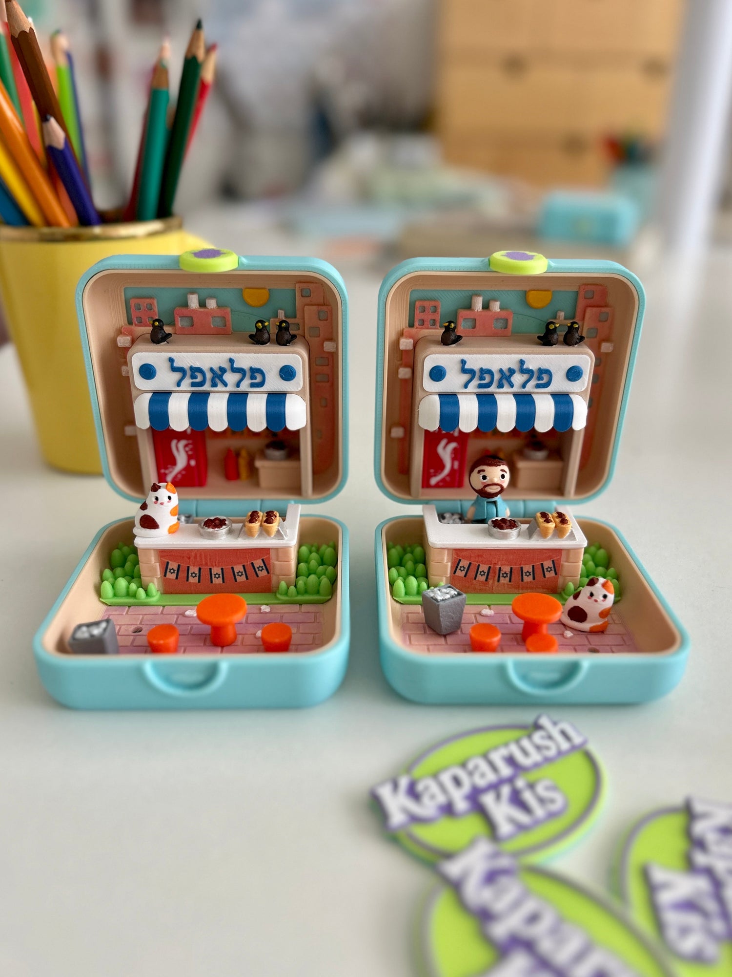 Miniature KaparushKis toy sets with tiny Israeli street scenes inside collectible boxes, alongside custom hand-drawn pet portrait miniatures made from photos